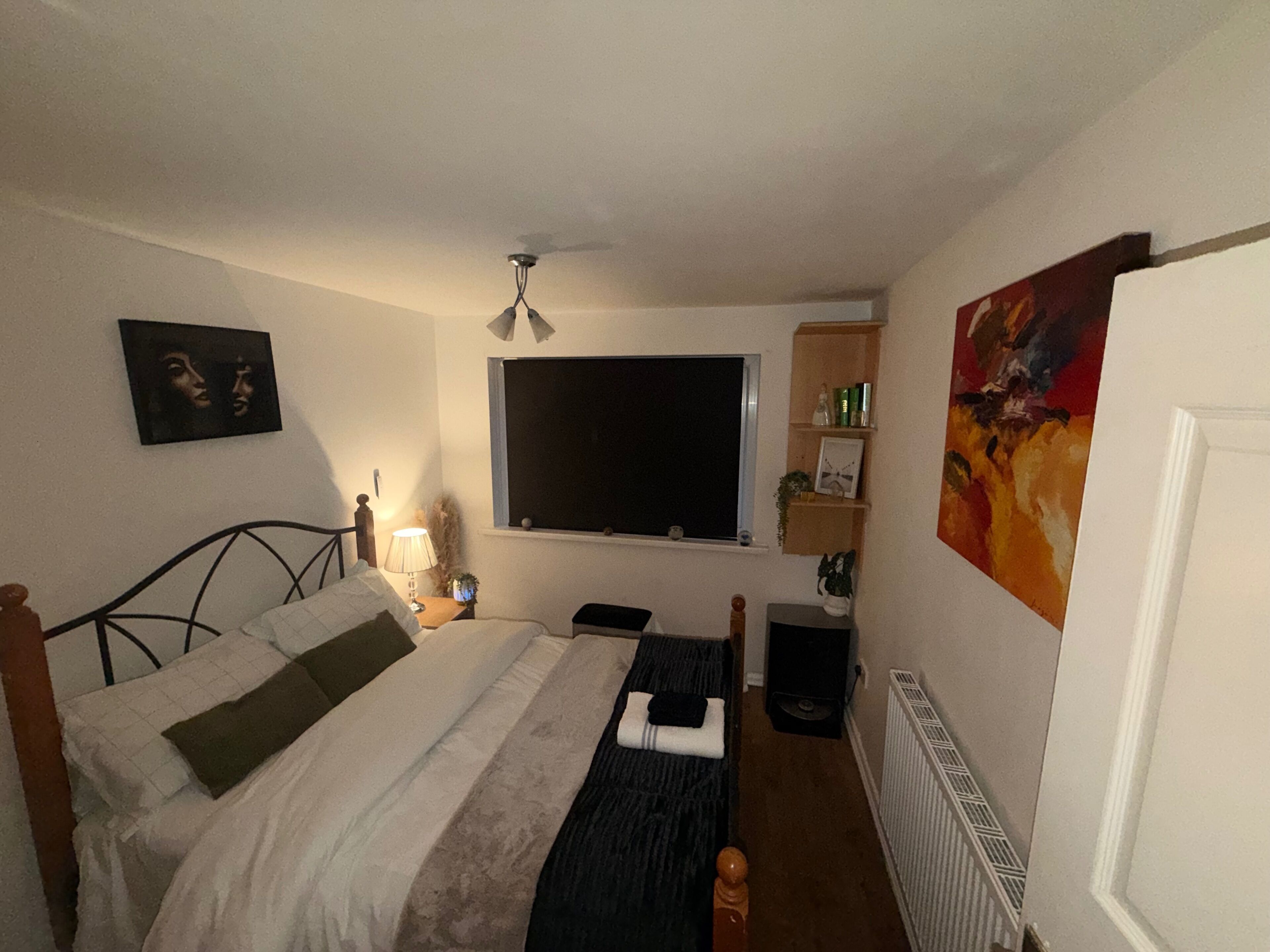 A bedroom with a bed, a bedside table with a lamp, a window, a radiator, and a painting on the wall.