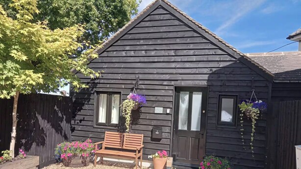 A wooden house with a front bench, hanging flower baskets, and a door with a window. - The Barn, Enstone - a Cosy Cotswolds Retreat (Chipping Norton)