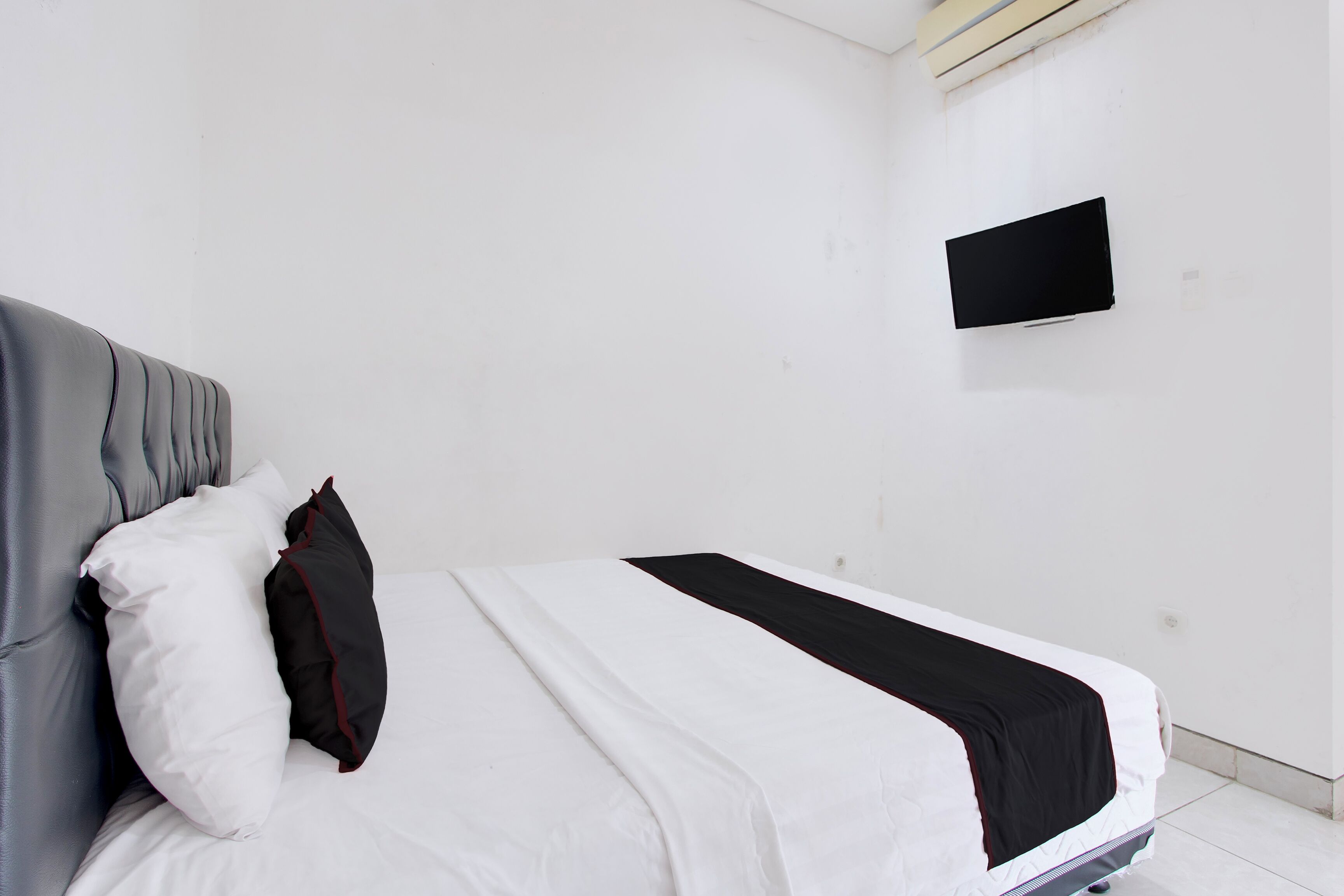 A bed with white and black bedding, a grey upholstered headboard, and a flat-screen TV mounted on the wall.