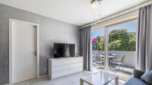 Ein modernes Wohnzimmer mit Flachbildfernseher, Glastisch, Sofa und einer Schiebetür zu einer Außenpatio mit Tisch und Stühlen.