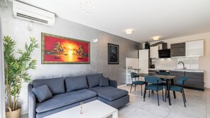 Ein modernes Wohnzimmer mit einem grauen Ecksofa, einem weißen Couchtisch und sesseln in Türkis. Im Hintergrund ist eine Küche mit grauer Arbeitsplatte und Edelstahlgeräten zu sehen.