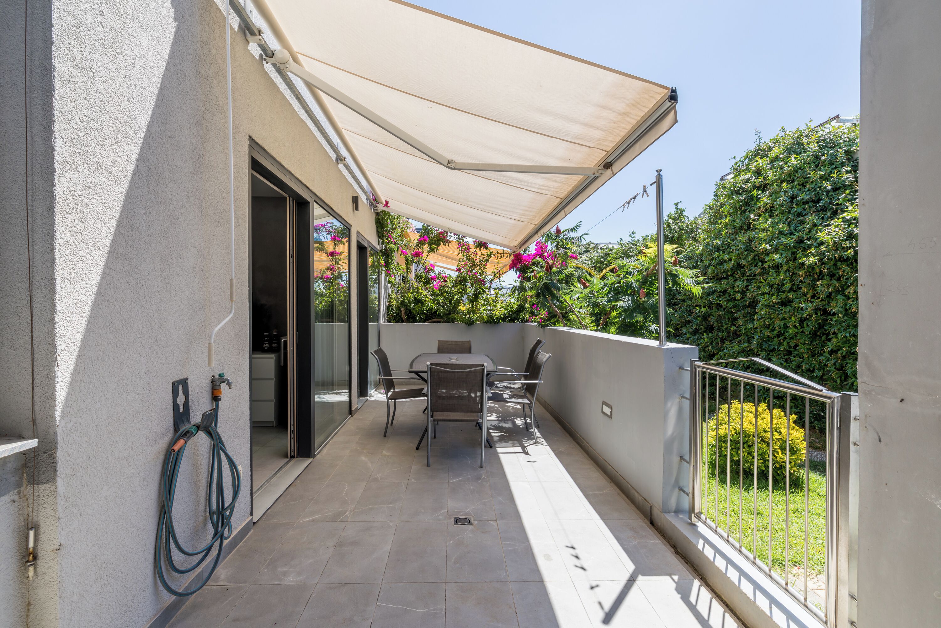 A covered patio with a table and chairs, a hose, and a view of greenery.