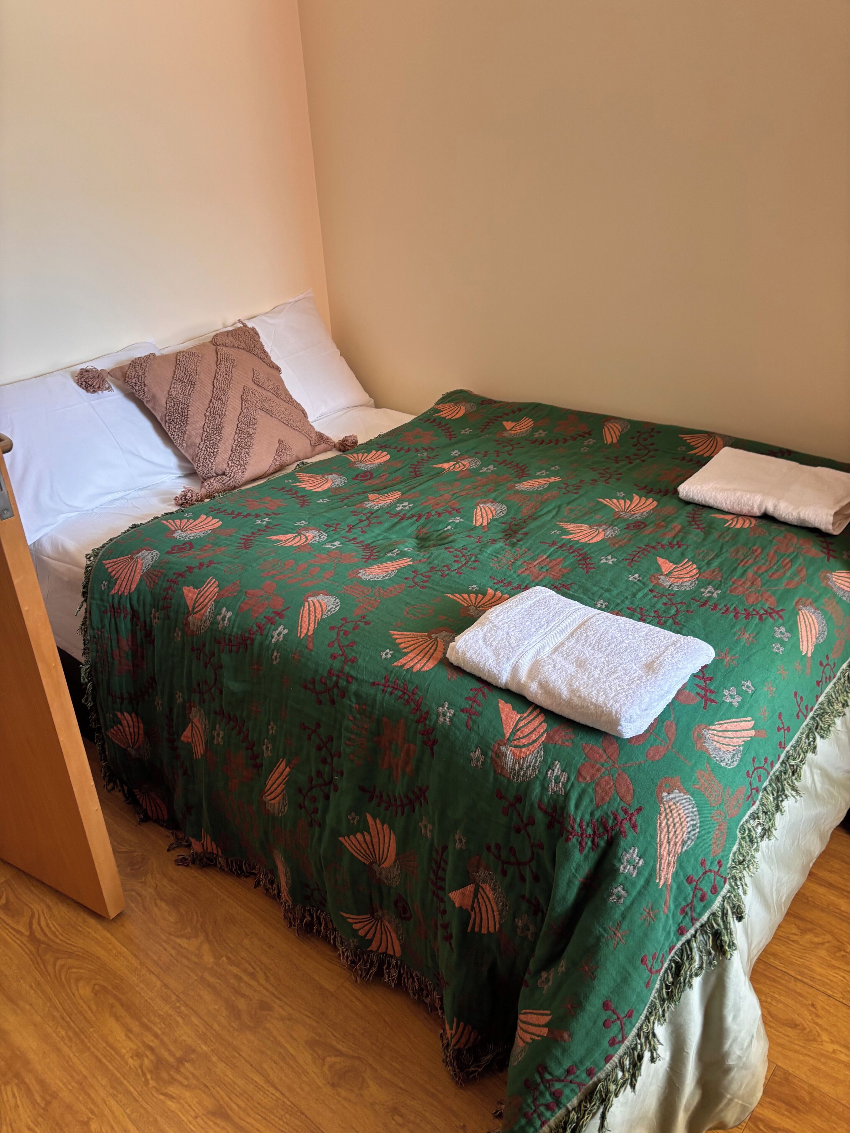 Un lit bien fait, recouvert d’une couvre-lit à motifs floraux verts, avec un oreiller blanc et une couverture marron.