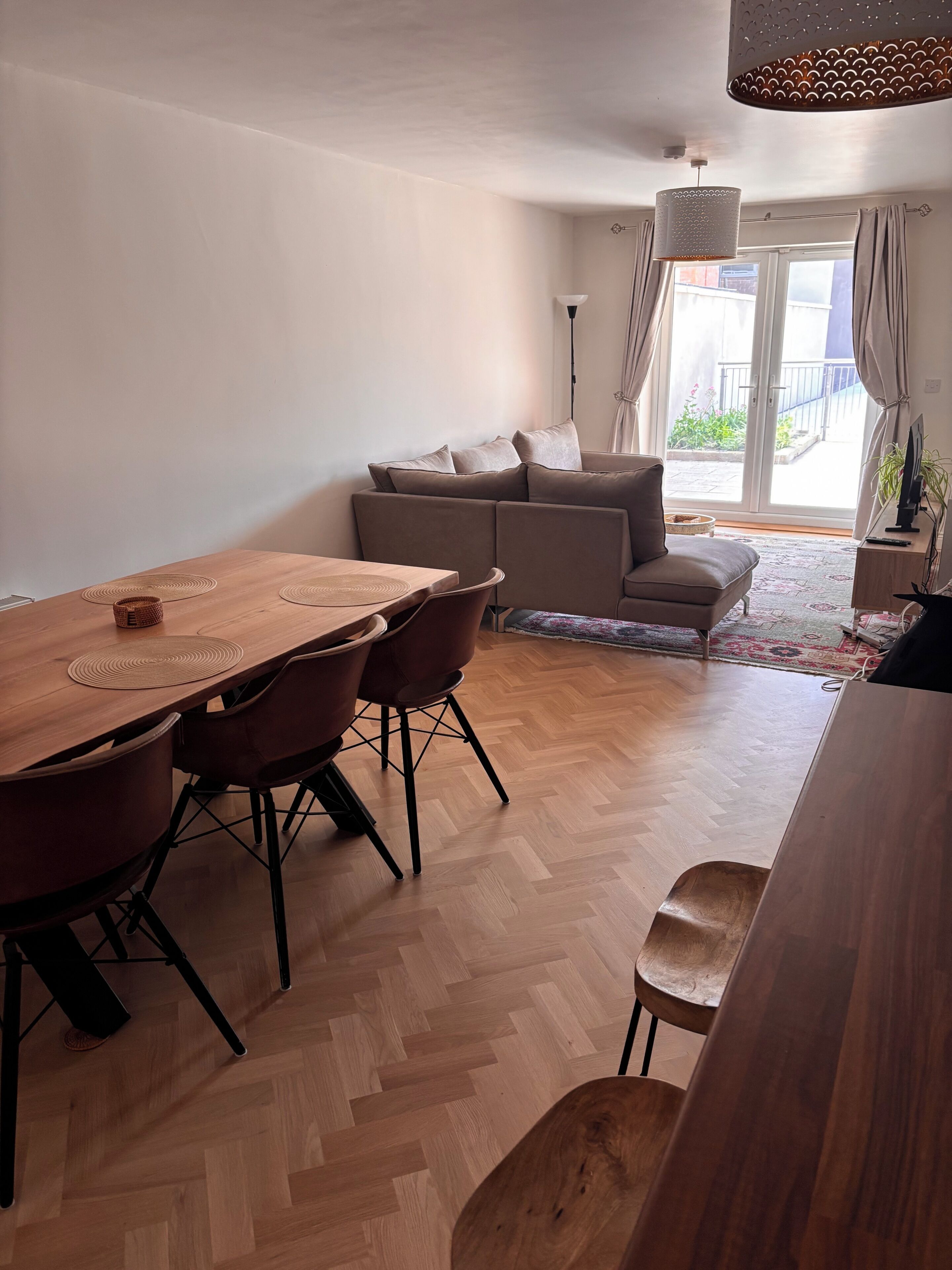 A modern living room with a wooden dining table, sofa, and a large window with curtains.
