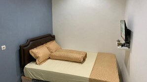 Una cama individual con cabecera, una mesita de noche con lámpara, y un estante en la pared con una planta.