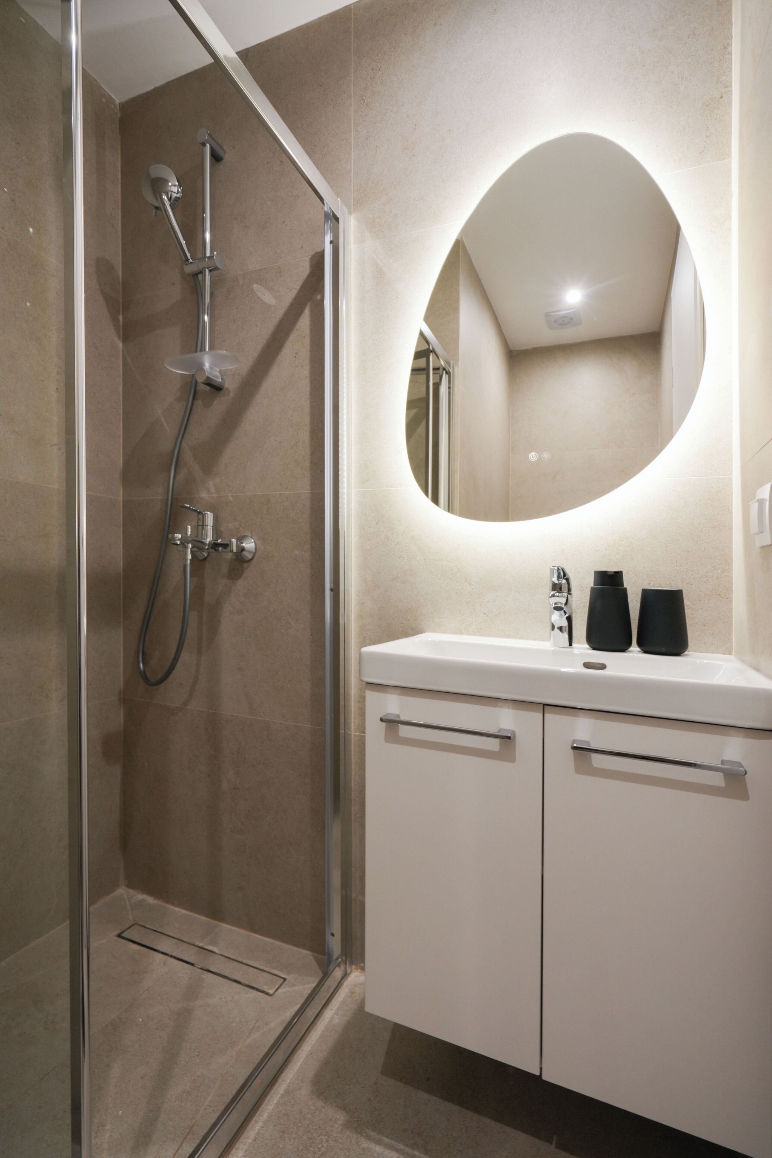 A modern bathroom with a glass-enclosed shower, a white vanity with a round mirror, and a beige tiled wall.