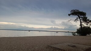 A beach with sand, a tree, and calm water with boats. - New apartment in a luxury residence on the waterfront of Lake Carcans. (Carcans)