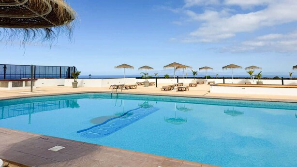 En swimmingpool med solstolar och parasoller, en byggnad med halmtak och utsikt över havet.