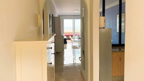 En hall med en välvd dörröppning som leder till ett matområde, ett köksområde med skåp och utsikt över stranden.
