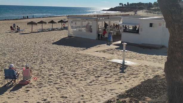 Uma praia com espreguiçadeiras, guarda-sóis, um pequeno quiosque e um edifício branco com telhado plano.