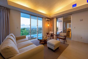 Ein geräumiges Wohnzimmer mit einer Couch, einem Hocker und einem Glastisch. Große Schiebeglasfenster führen zu einem Balkon mit Blick auf Grünflächen und einen Pool.