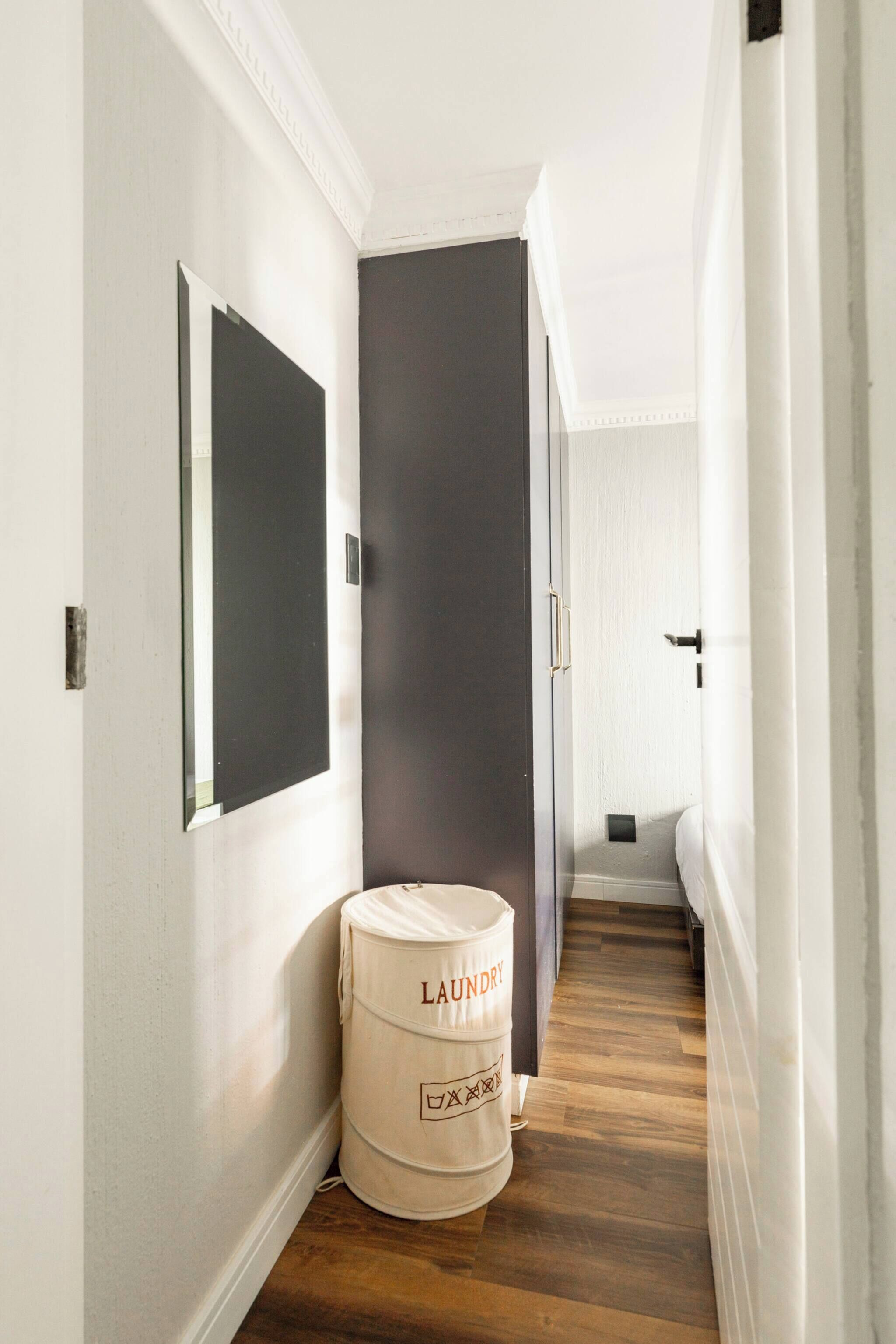A narrow hallway with a dark door, a white wall with a mirror, and a laundry barrel.