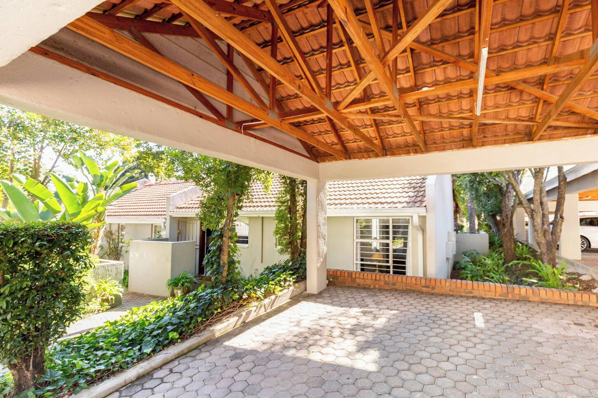 A covered patio area with a tiled floor, surrounded by greenery and a building with a tiled roof.