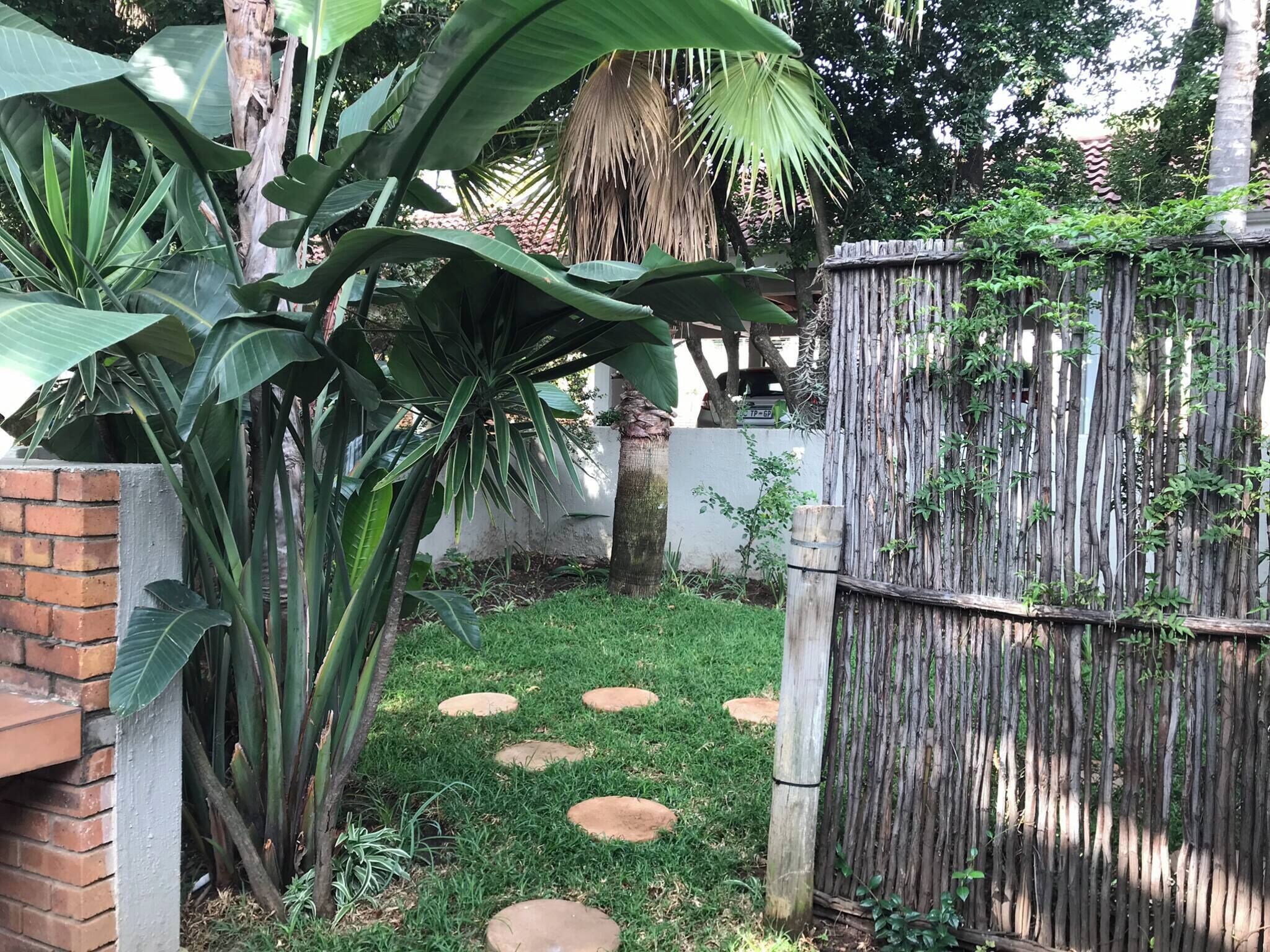 Een tuin met een bakstenen muur, grote groene planten, een houten omheining en staptegels.