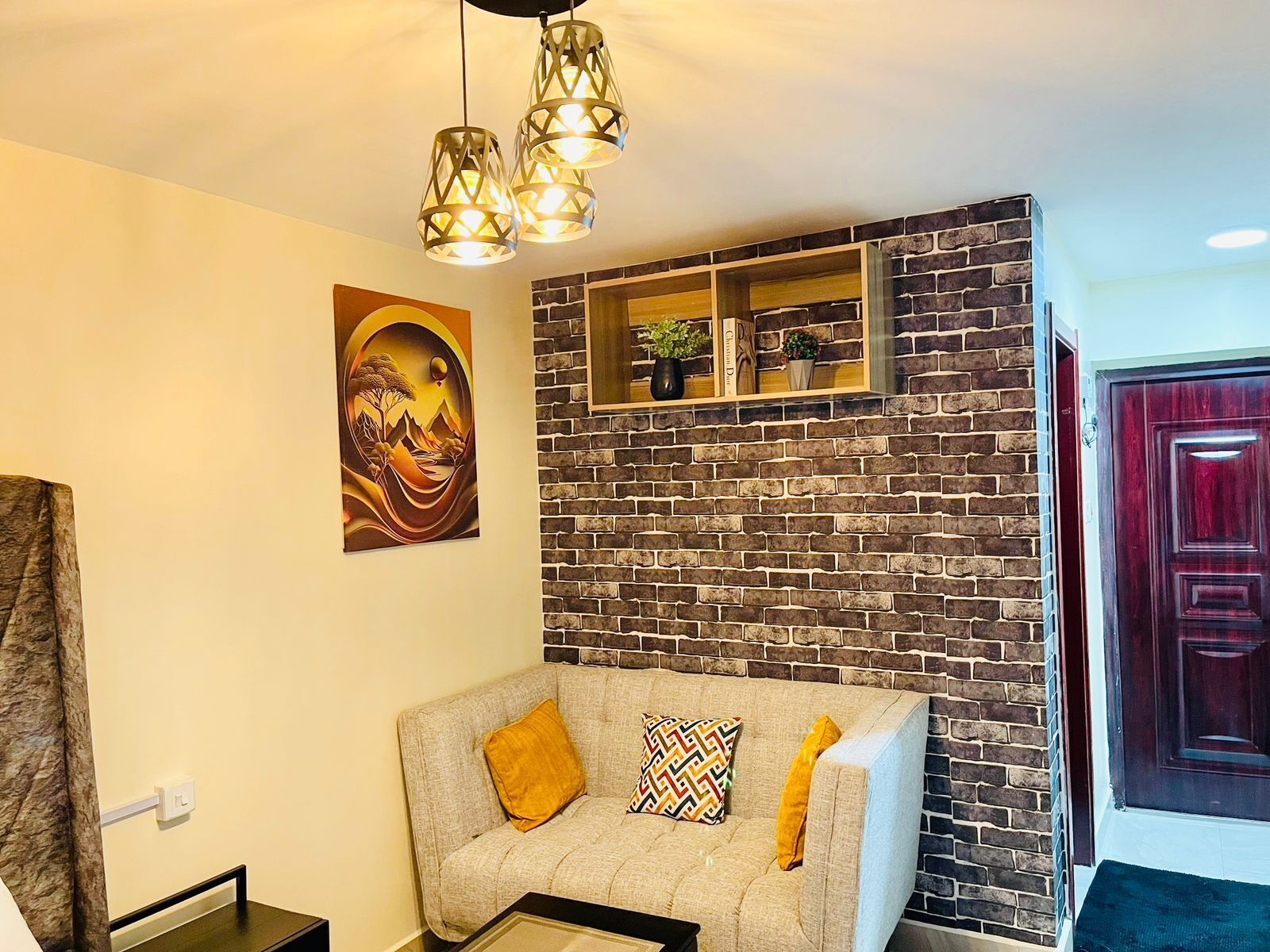 A living room with a brick wall, a sofa with cushions, a painting, and a wooden shelf with decorative items.