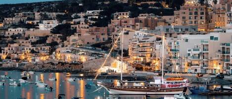 A coastal town at dusk with a marina filled with boats, buildings with balconies, and a prominent hill in the background.