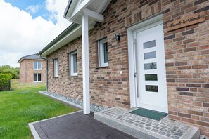 A brick building with a white door, a small porch, and a sign above the door. - New Construction, 125 square meters | Hot Tub • Wallbox • Underfloor heating | freestanding | 6 people (Büsum)