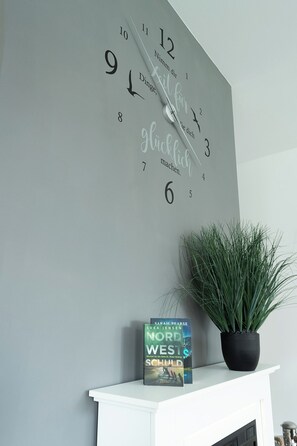 Eine Wanduhr mit deutscher Schrift, ein Buch mit dem Titel 'Nord West SCHULD' und ein Topfpflanze auf einem weißen Kaminbrett.