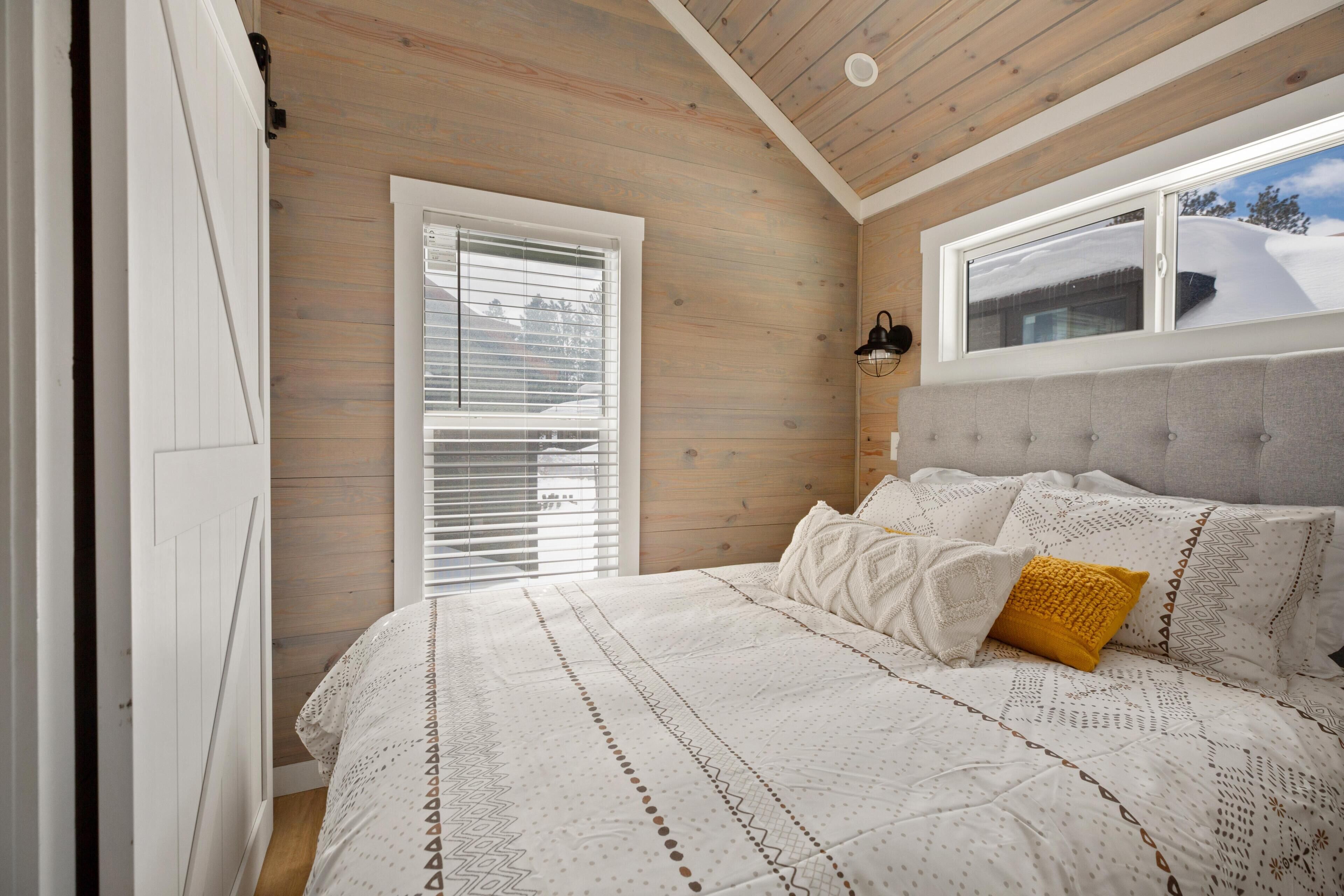 Quarto com teto de madeira, cama grande com roupa de cama estampada, janela com persianas e vista para uma paisagem nevada.
