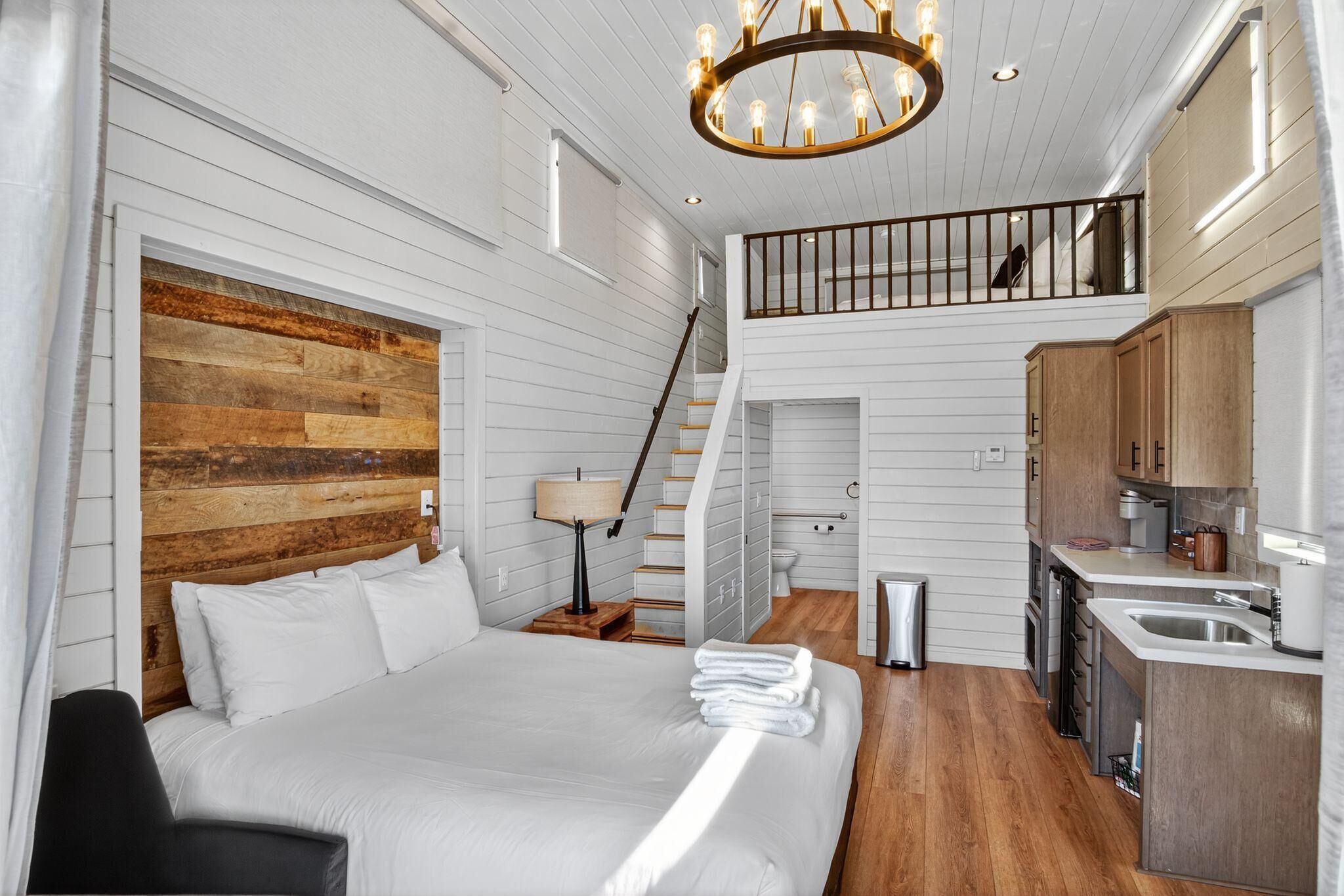 A bedroom with a large bed, a wooden accent wall, a staircase to a loft, and a kitchenette.