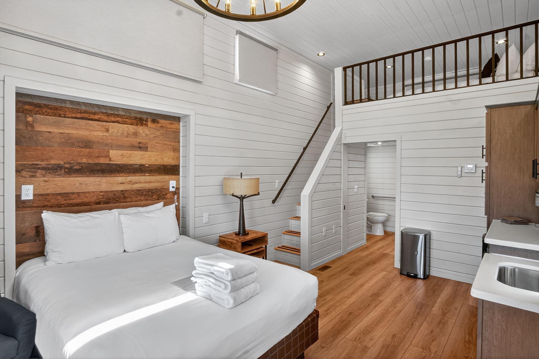 Ein Schlafzimmer mit hölzernem Kopfteil, einem Bett mit weißer Bettwäsche, einem Nachttisch mit Lampe, einer Treppe zu einer oberen Ebene sowie einem kleinen Badezimmer mit Toilette und Waschbecken.