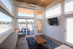 Quarto com sofá, mesa e televisão. Grandes janelas e porta que dá para o exterior.