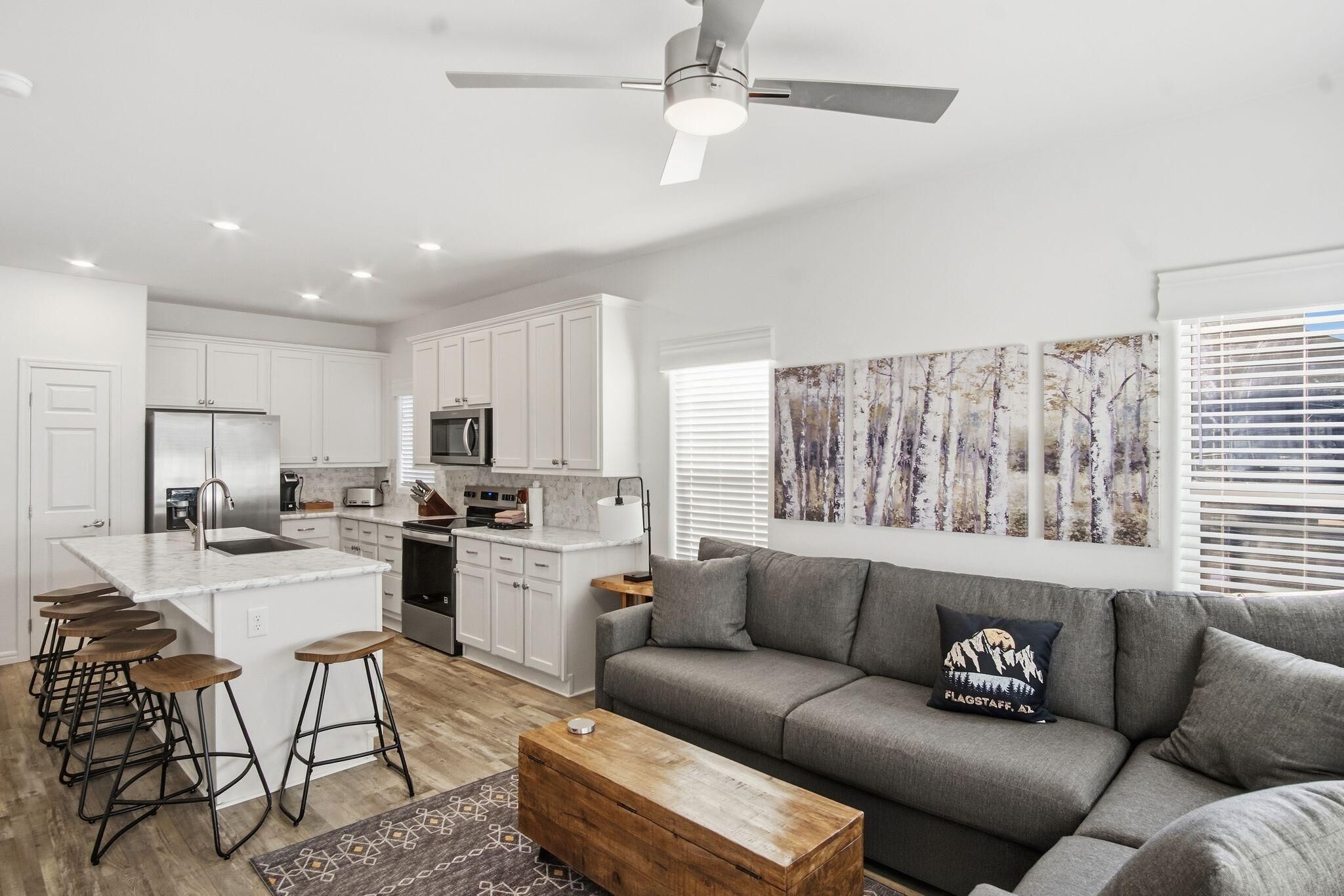 A modern kitchen with white cabinets, a central island with stools, a grey sofa with a decorative pillow, and a ceiling fan.
