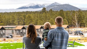 Una familia de tres personas contemplando un paisaje montañoso con una hilera de cabañas al fondo.