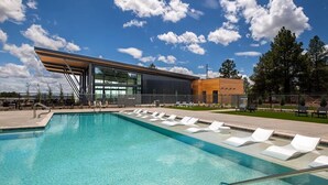 Un área de piscina moderna con agua azul cristalina, sillas de descanso y un edificio con grandes ventanales y techo inclinado.