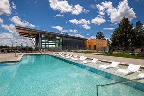 Un espace piscine moderne avec une eau d’un bleu limpide, des chaises longues et un bâtiment doté de grandes fenêtres et d’un toit incliné.