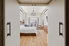 Une chambre à coucher avec un lit, un lustre, un bureau et une vue sur l’extérieur, visible à travers une porte.