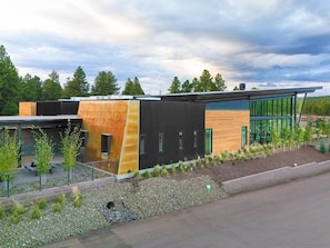 Een modern gebouw met een combinatie van houten en donkere panelen, een grindpad met jonge bomen, en een heldere lucht.