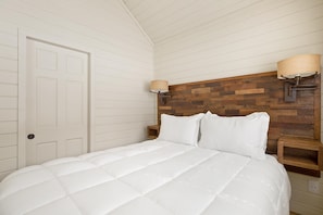Quarto com cama branca, cabeceira de madeira, duas mesas de cabeceira e porta branca.