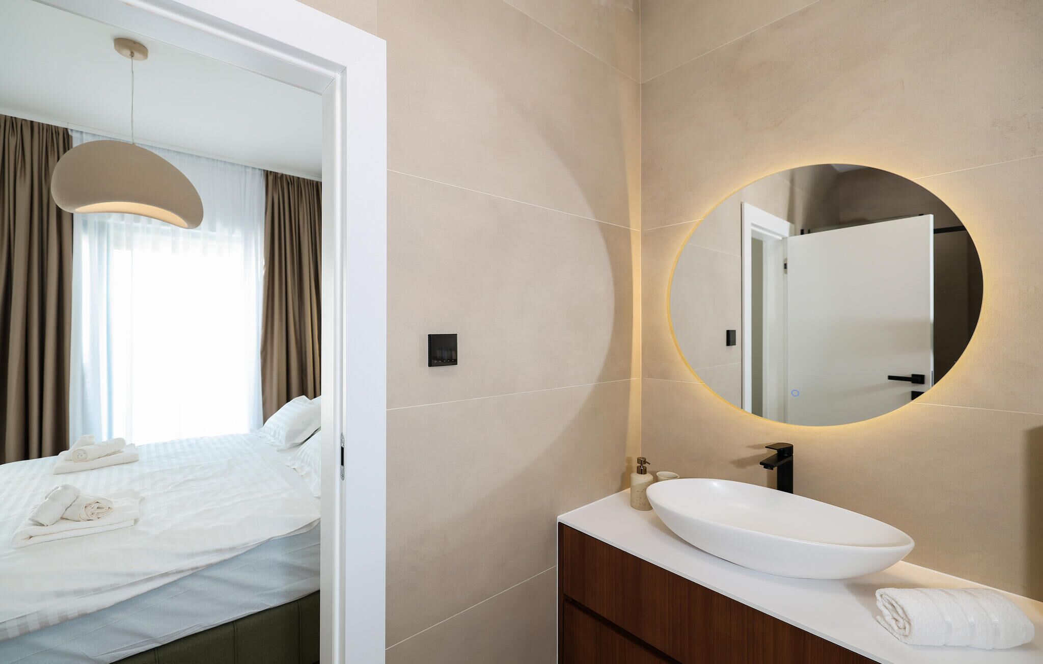 Ein modernes Hotelzimmer mit Bett, rundem Spiegel, weißem Waschbecken und Blick ins Zimmerinnere.
