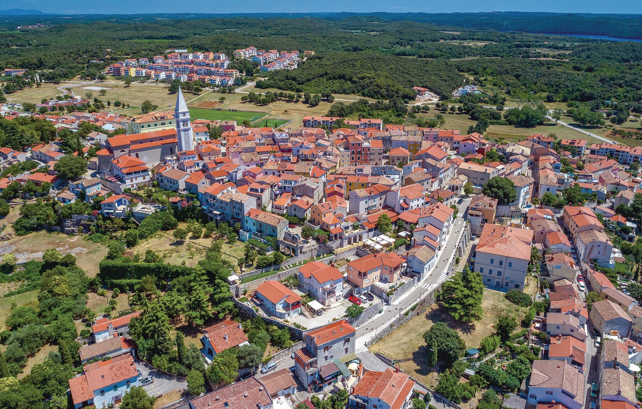 An aerial view of a densely packed town with red-tiled roofs, a prominent church tower, and a mix of residential and commercial buildings.