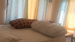 A bedroom with a bed, pillows, curtains, a small table, and a patterned rug. - We are not a commercial hotel... we are your home (Munsyari)