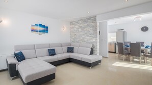 Ein modernes Wohnzimmer mit einer Eckcouch, einem Essbereich mit Stühlen, einer Küche im Hintergrund und einer Wand mit Steinverkleidung.