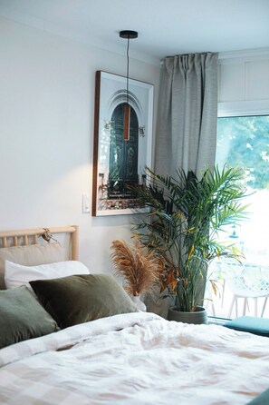 Ein Schlafzimmer mit einem Bett, einer Topfpflanze, einem Wandbild und einem Fenster mit Vorhängen.