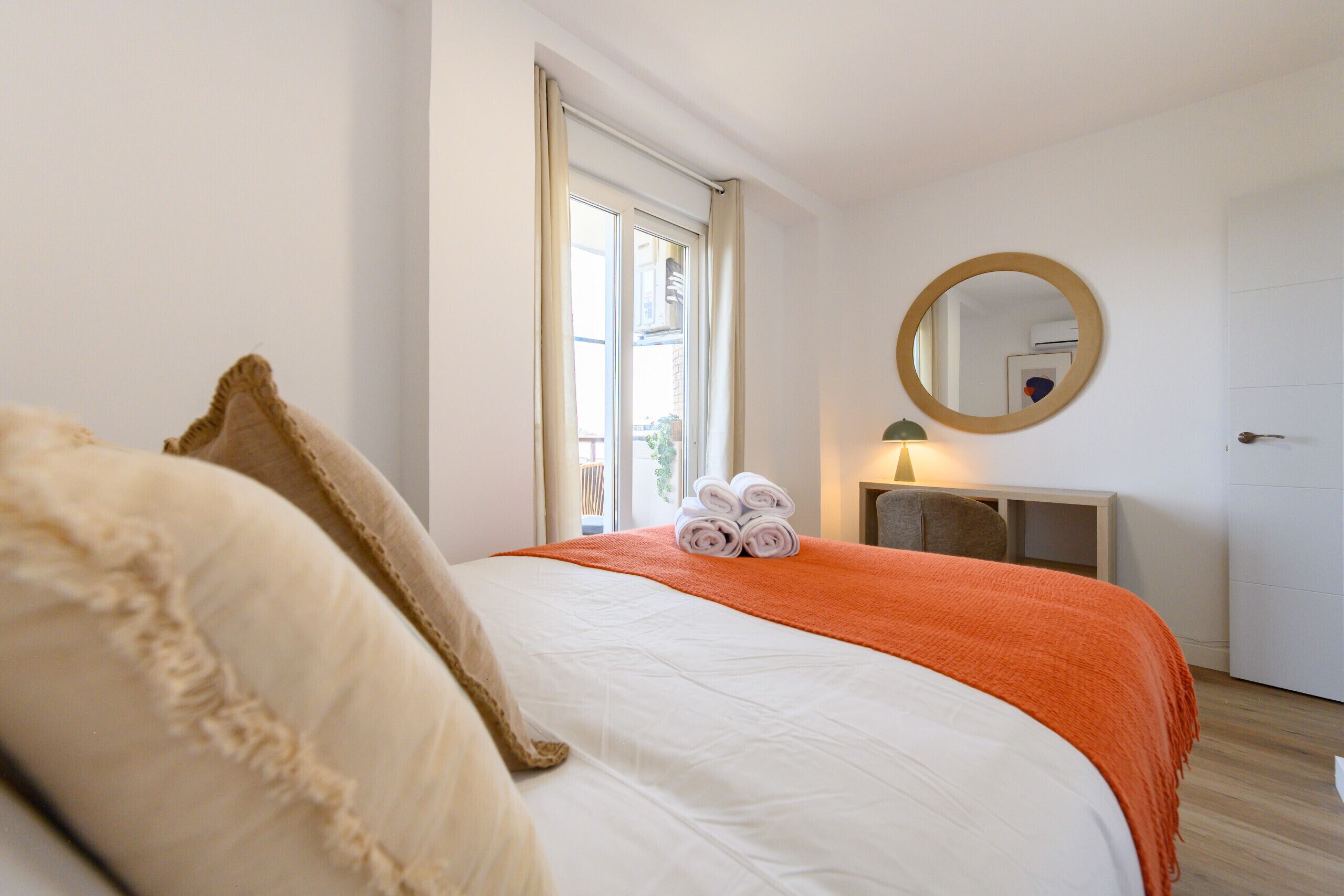 Una camera d'albergo con un letto, uno specchio rotondo, un comodino con una lampada e una finestra con tende.