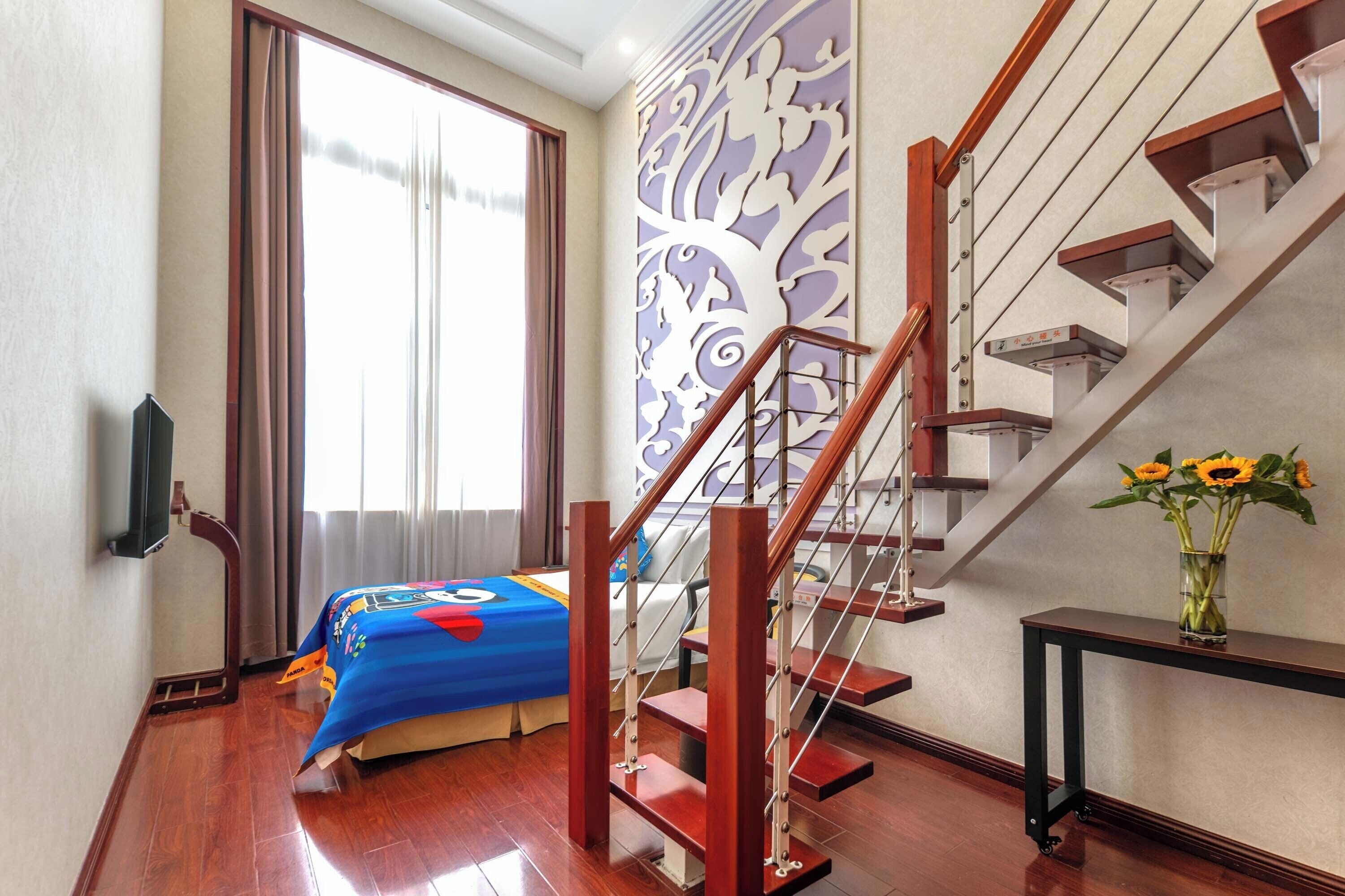 Una camera d'albergo con una scala in legno, un letto con copriletto blu, una grande finestra con tende e una parete con un pannello decorativo.