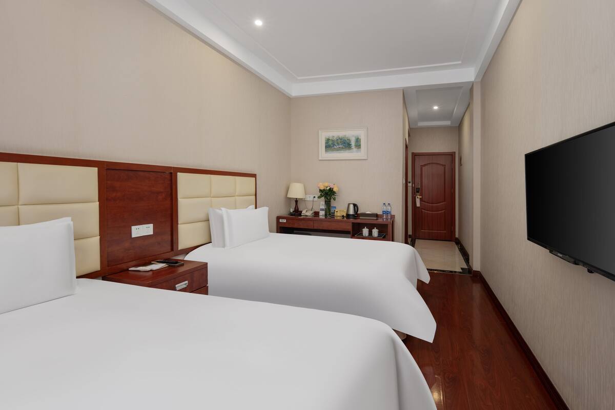 A hotel room with two beds, a desk with a lamp, a television, and a painting on the wall.