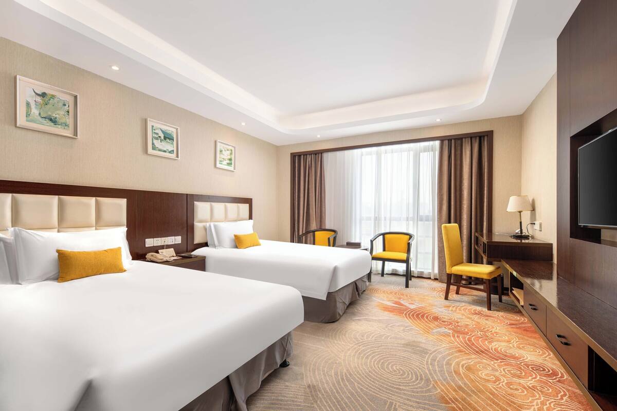 A hotel room with two beds, a desk, a chair, a TV, and a large window with curtains.