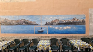 Um mural retrata uma vila costeira com um pequeno barco, uma mesa com toalha estampada e várias cadeiras de plástico.