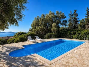 En rektangulär swimmingpool med en kant av sten, två solstolar och utsikt över havet och bergen.