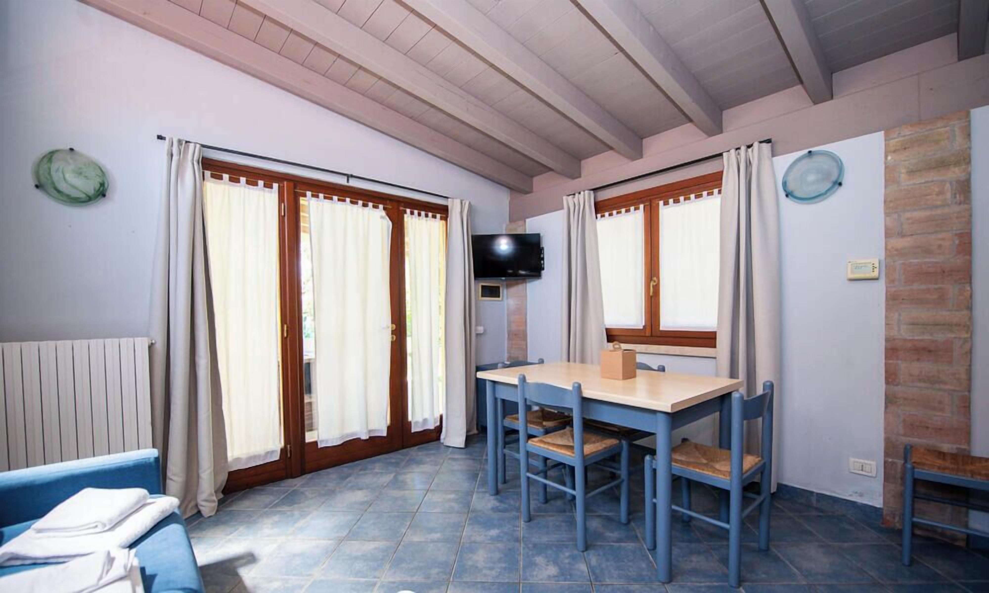 A room with a blue tiled floor, a wooden table with chairs, a television on the wall, and a window with curtains.