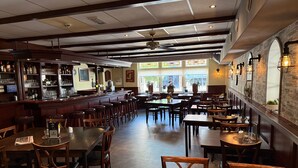Un interior de restaurante con mesas y sillas de madera, una zona de bar y ventanas amplias que permiten la entrada de luz natural.