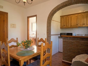 A dining area with a wooden table, chairs, and a vase of oranges. There is a kitchen with wooden cabinets and a brick archway. - Las Rosas 5 by Casasol (Frigiliana)
