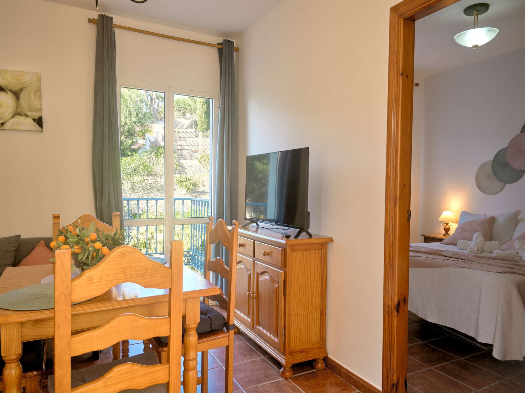 Une pièce comprenant une table à manger en bois, des chaises, un téléviseur posé sur un meuble en bois, un lit recouvert d’une courtepointe blanche et une vue sur l’extérieur à travers une fenêtre ornée de rideaux.