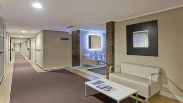Un vestíbulo de hotel con mostrador de recepción, un área de espera con sofá y una mesa de centro, y un pasillo que da a las habitaciones.