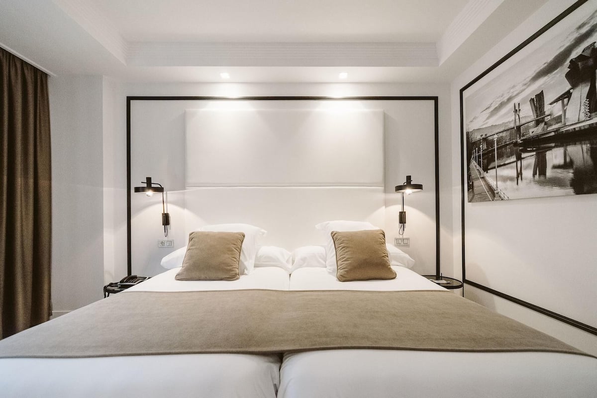 Una habitación de hotel moderna con una cama grande, dos lámparas de noche, un cabecero y un cuadro enmarcado en la pared.